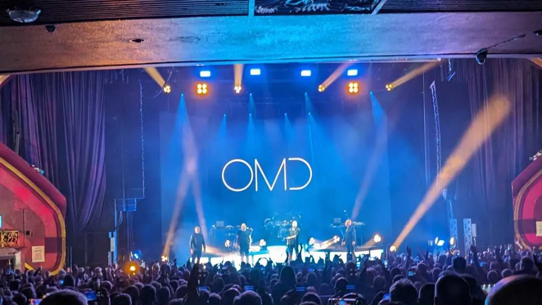 A concert in a big hall with the band name, OMD, written on the wall behind the stage