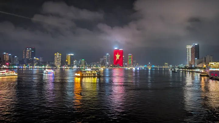A night time view taken of Da Nang from a bridge. A building is lit up with the Vietnamese flag