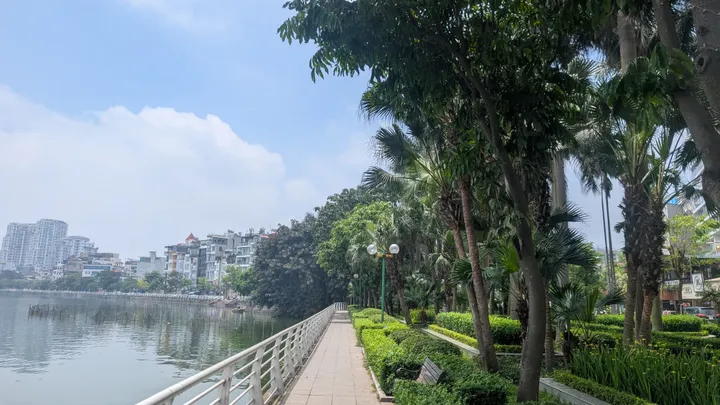 A path with a lake to the left over a railing. Trees to the right and buildings in the background