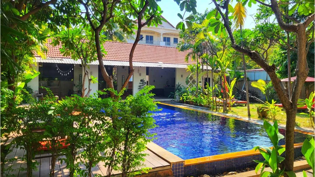 A villa with a pool and trees surrounding it, with a building in the backgronud