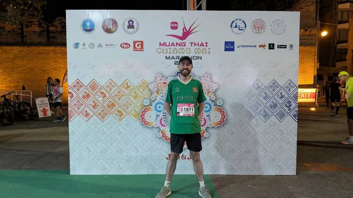 A man in a green t-shirt and shorts poses in front of a sign for the Chiang Mai Marathon