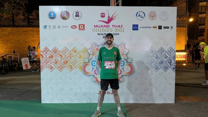 A man in a green t-shirt and shorts poses in front of a sign for the Chiang Mai Marathon